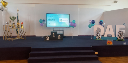 A decorated stage for the Dar Al Fikr Sports Olympics 2025 featuring trophies, a winners’ podium (1–2–3), school-colored balloons, a display screen, and large DAF letters decorated with soccer balls.